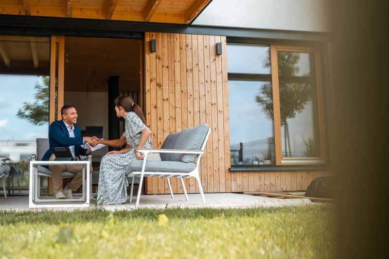 A Hispanic real estate agent finalizes a transaction with a smiling diverse couple, shaking hands on a sunny patio, symbolizing successful homeownership.