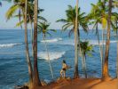A person walks on a sandy beach path toward the ocean under a blue sky.