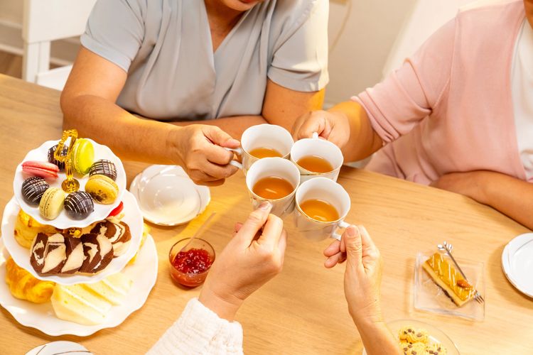 Four people cheers with tea cups at a table with assorted pastries and desserts.