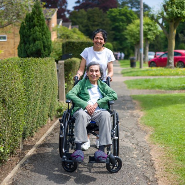 A young woman joyfully pushing an elderly woman in a wheelchair along a sunny park path.