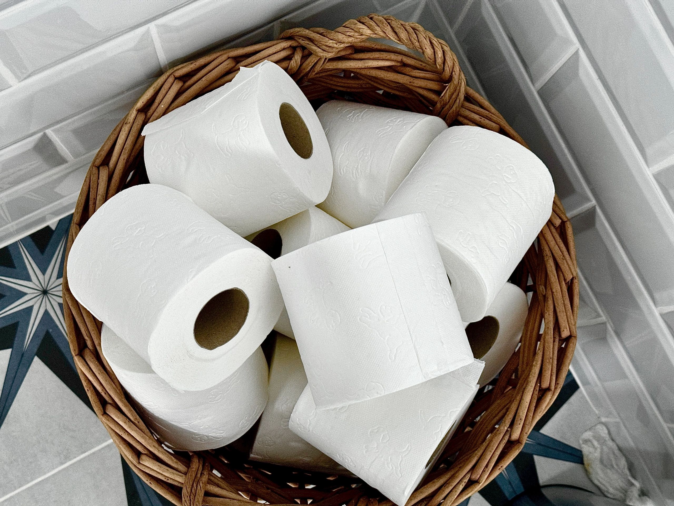 A basket filled with multiple white toilet paper rolls in a tiled bathroom corner.