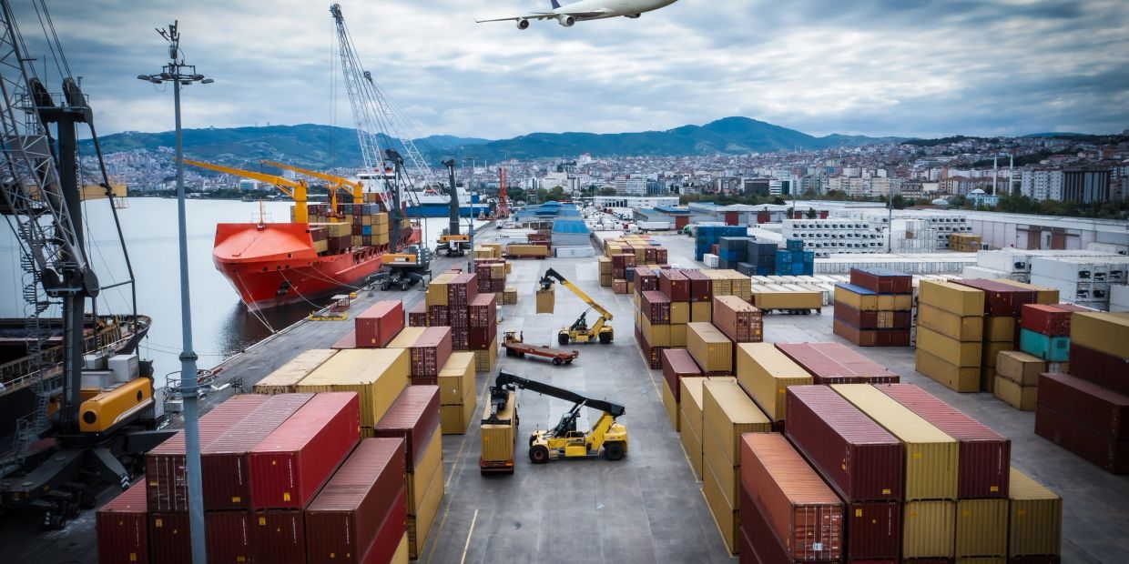 Busy shipping port with containers, cranes, and a plane overhead.