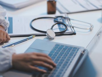 Healthcare professional typing on a laptop with a stethoscope nearby.