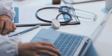A person typing on a laptop with stethoscope and papers.