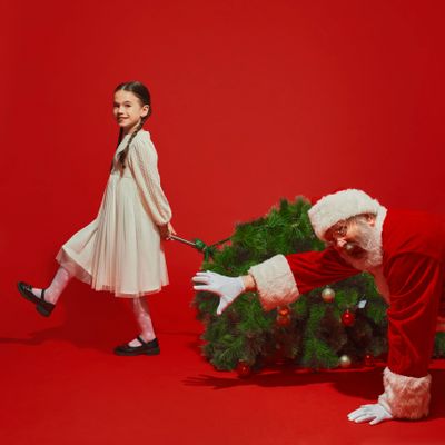 Girl playfully pulling a Christmas tree with Santa crawling behind it on red background.