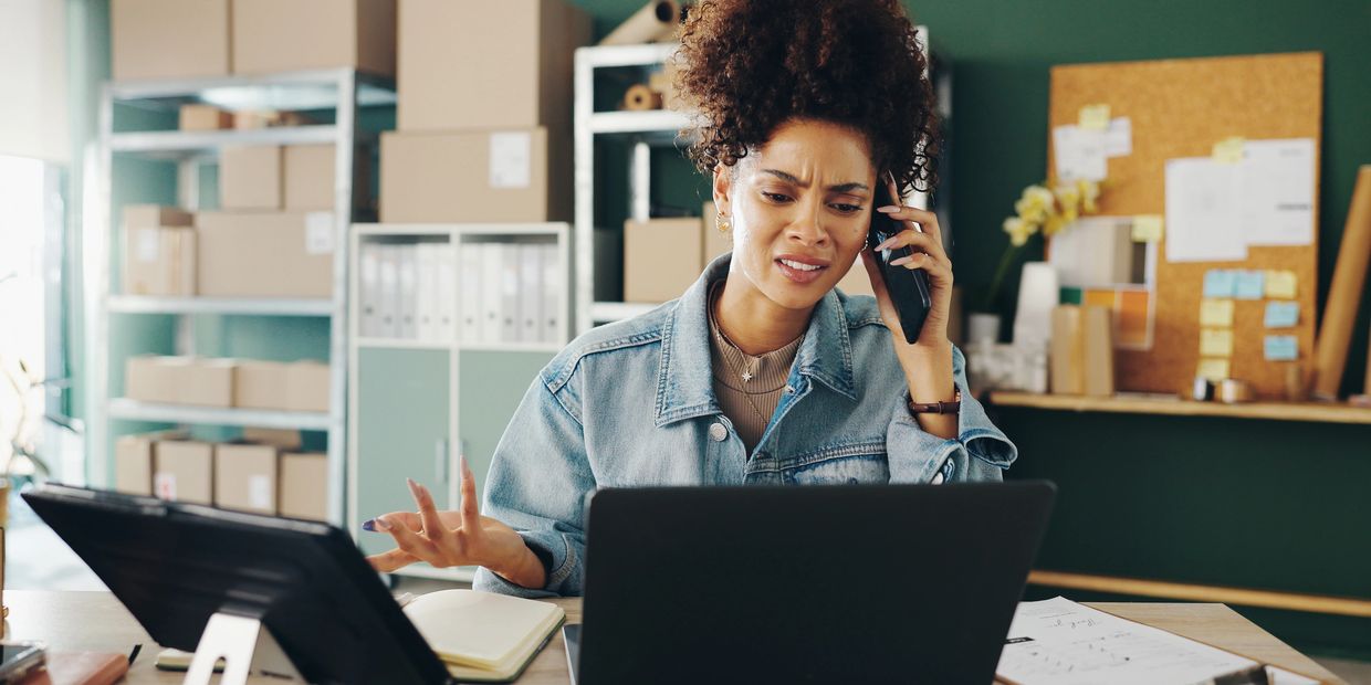 Frustrated woman on phone, working on laptop in a cluttered office.