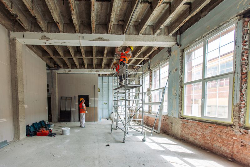 Image of construction workers on scaffolding, wearing safety helmets and vests, engaged in the renovation of an old building with exposed brick and large windows.