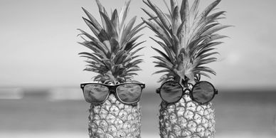 Two pineapples wearing colorful sunglasses on a sunny beach.