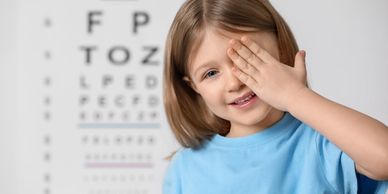 A young girl covering one eye during an eye exam with an eye chart in the background.