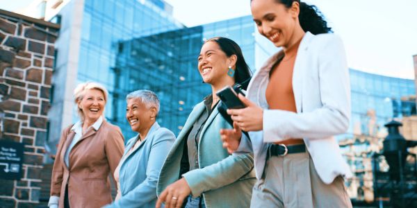 Four women in business attire walking and smiling outdoors.