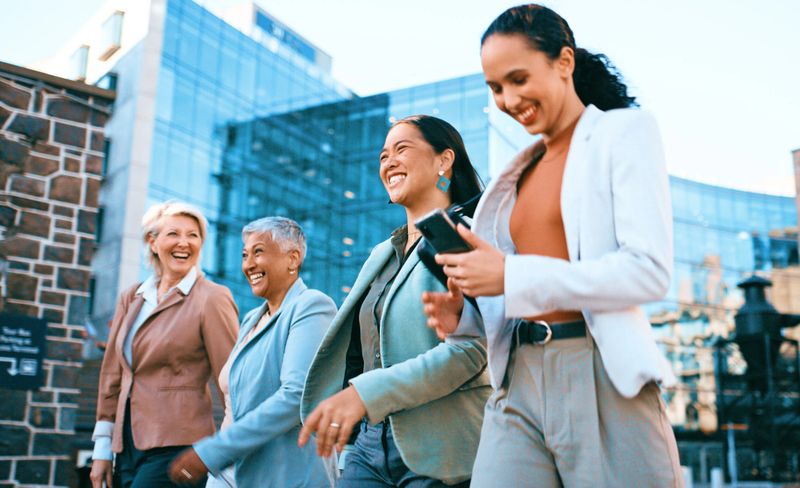Diverse group of professional women walking, smiling, and using a smartphone outdoors in a modern business district.