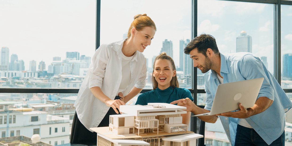 Three architects discuss a modern building model in a bright office with city views.
