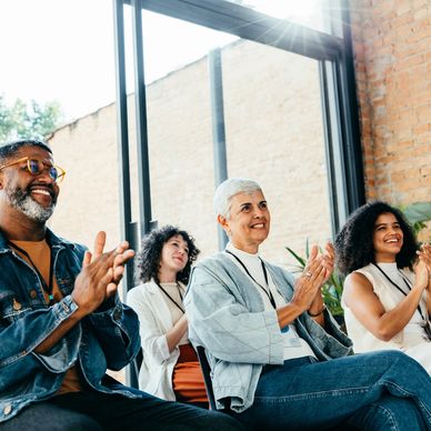 A group of diverse people clapping and smiling during an indoor event.