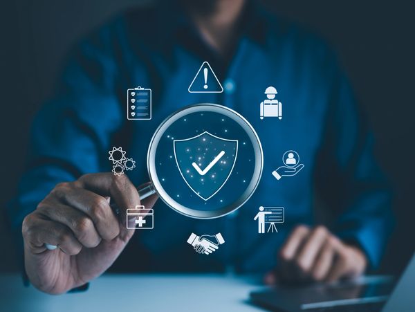 Person holding magnifying glass over shield with checkmark and security icons.