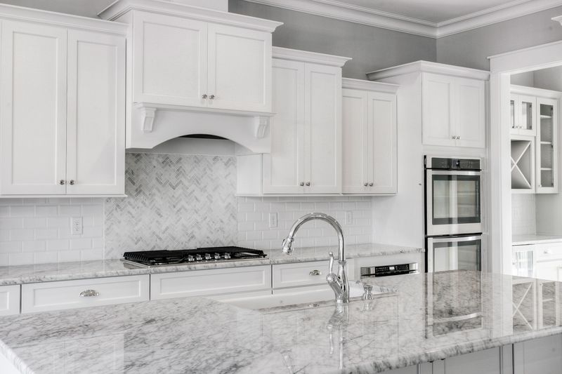 A sleek, modern kitchen featuring white cabinetry, marble countertops, a gas range, and stainless steel fixtures. The herringbone tile backsplash adds a touch of elegance, while the spacious design and bright lighting create an inviting and functional cooking space.