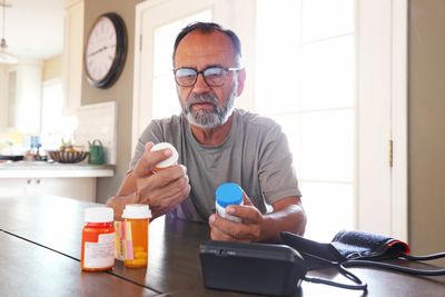 Elderly man reading medication labels at home with blood pressure monitor nearby.