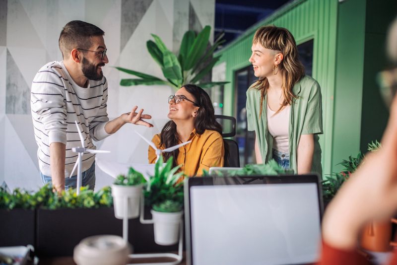 Office team having a discussion about an ongoing project, gathered around a desk with a laptop and surrounded by greenery, focusing on new ideas and effective strategies.