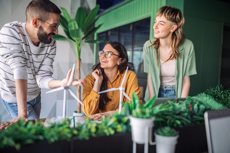Three young adults, two females and one male, actively brainstorm and discuss wind turbine designs together at a desk filled with plants in a modern, collaborative office space.