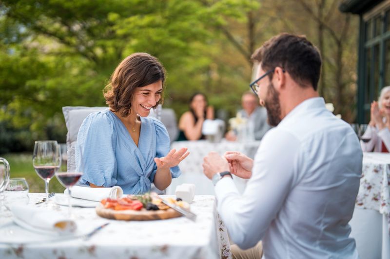 A mid-adult Latin male presents a ring to his elated Caucasian female partner during an intimate terrace dinner, signifying a romantic engagement, surrounded by elegant dining attire and soft evening light.