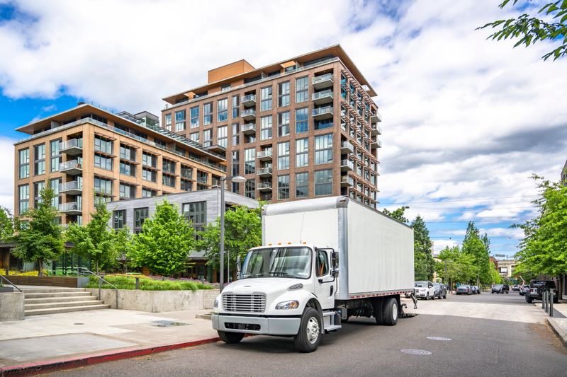 Middle duty mover day cab white compact bonnet rig semi truck with box trailer unloading delivered to customer apartments goods standing on the urban city street at summer cloudy day