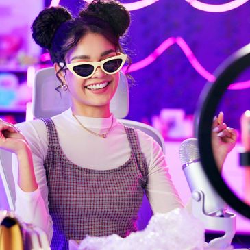 A young woman with sunglasses smiles while streaming with a microphone and ring light.