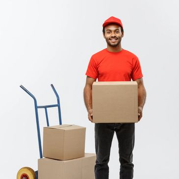On-demand delivery man in red uniform holding a cardboard box with a hand truck and boxes behind. Junk removal services being performed. 