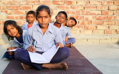 Children studying outdoors against a brick wall, focused on their notebooks.