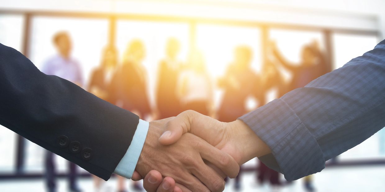 Two professionals shaking hands with blurred colleagues in the background.