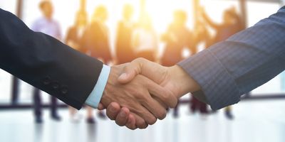 Two people shaking hands in a business setting with blurred colleagues in background.