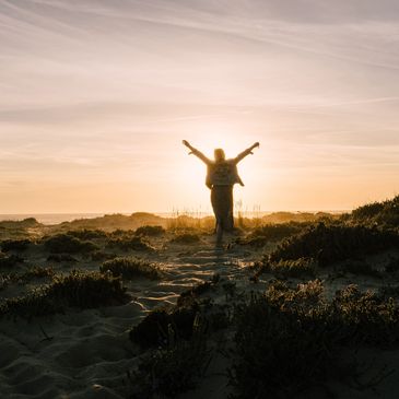 Person with arms raised enjoying a sunset on sandy dunes.