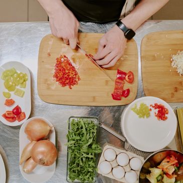 Person chopping red bell pepper on a wooden board surrounded by various fresh ingredients.