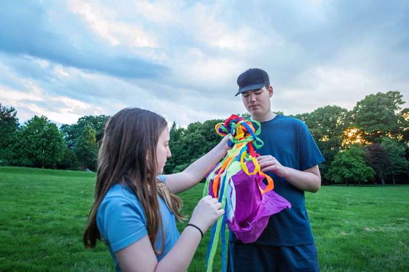 An autistic 18-year-old boy and his 10-year-old sister share a moment in a lush park during early summer evening. They are folding the kite together after playing with it. This activity unfolds against a backdrop of green grass and trees under a cloudy sky, highlighting sibling interaction and autism awareness.