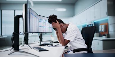 A stressed woman holding her head at a workstation with multiple monitors.
