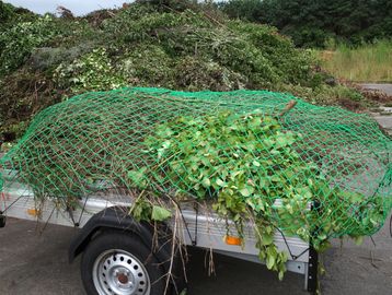 Trailer loaded with garden waste covered by a green net on a paved surface.