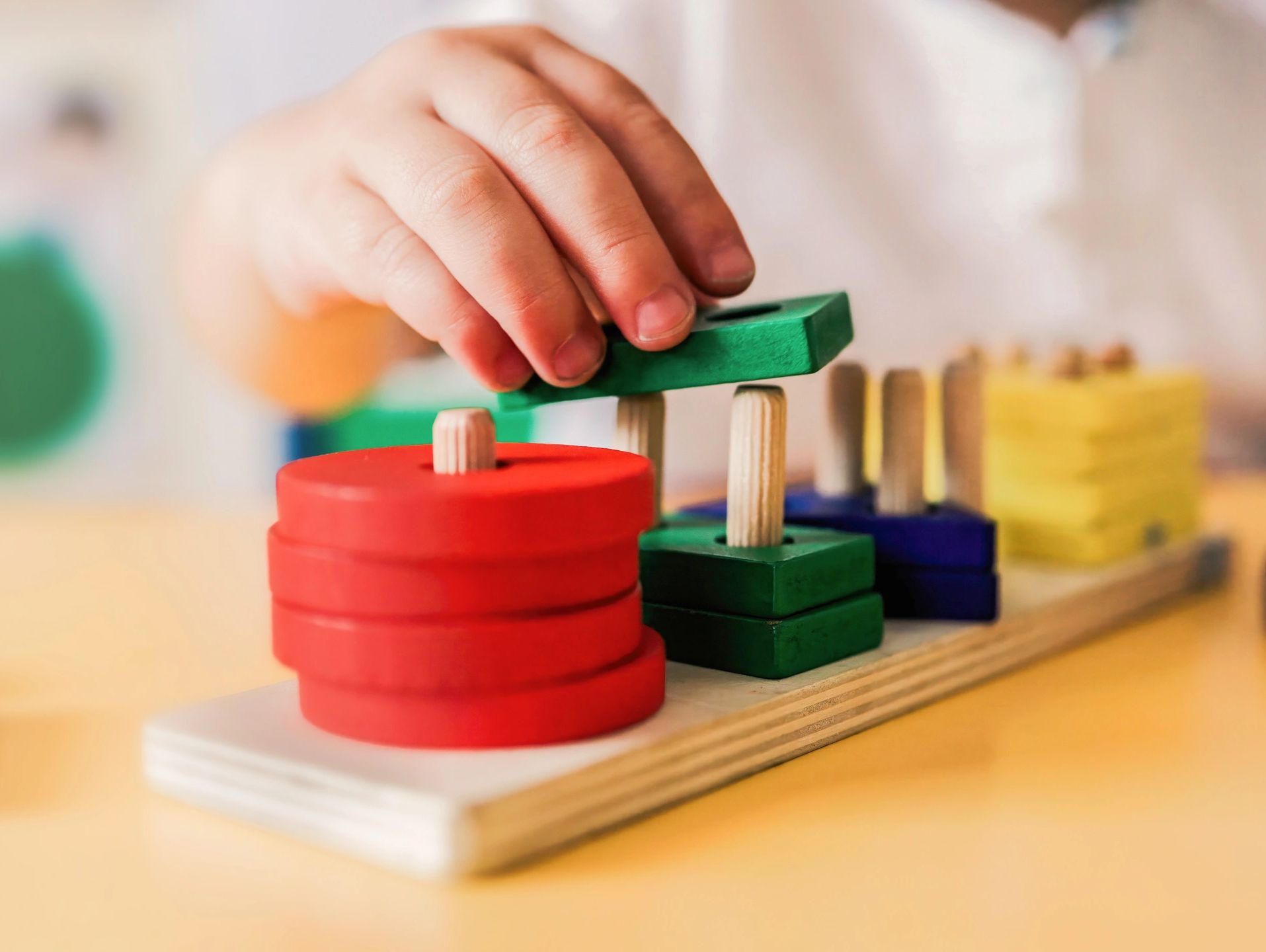 Child's hand stacking colorful wooden shapes on pegs for learning.