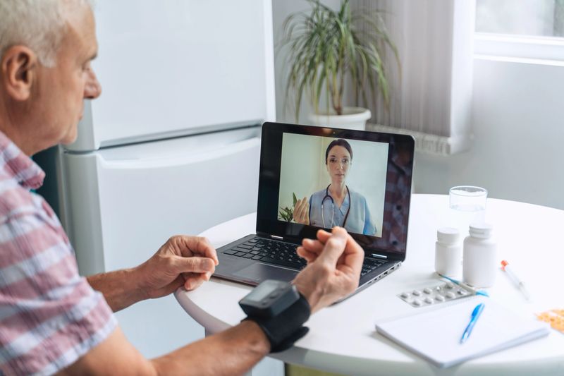 An elderly patient engages in an online conversation with a nurse, visible on his laptop screen, while keeping track of his blood pressure during a telehealth appointment.