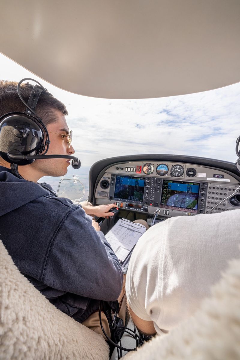 Pilot and co-pilot in the cockpit of a light aircraft, navigating with flight instruments and charts. The pilots wear headsets, and the view outside shows a cloudy sky.