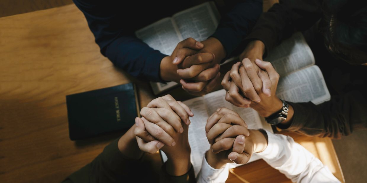 Four people holding hands in prayer over open Bibles on a wooden table.