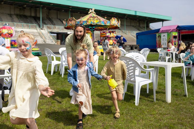 Family visiting an Easter fair in the North East of England. The father is chasing his three children, having fun with them on a sunny day. There are fairground rides visible behind them.