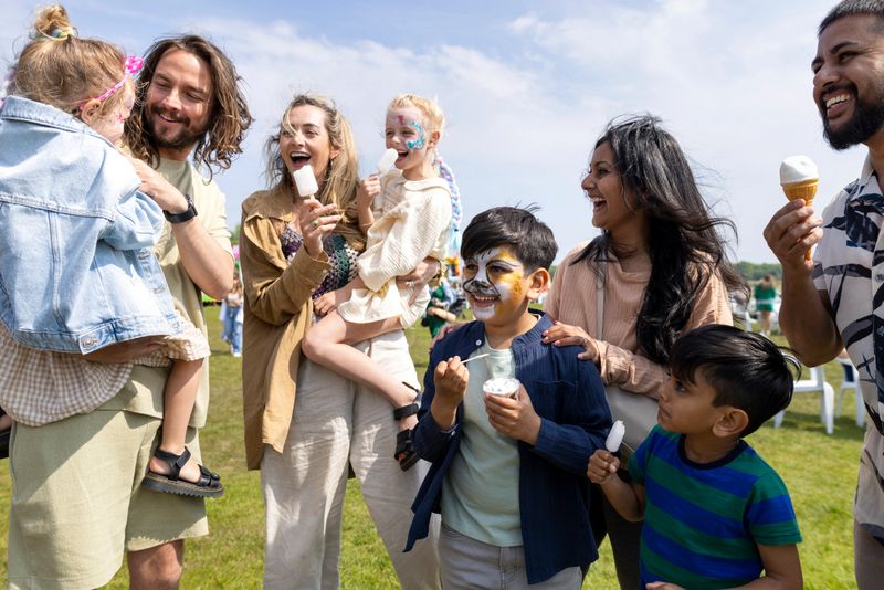 Two families visiting an Easter fair in the North East of England. They are standing together, eating ice creams and frozen lollies, enjoying the sweet treats while talking together. Some of the children have their faces painted.