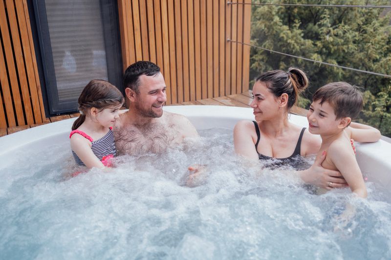 Happy family with two small children have fun in the hot tub. Cheerful young dad splashes water on his laughing cute son. Smiling man, woman, funny son and cute daughter relaxing in bubbling water