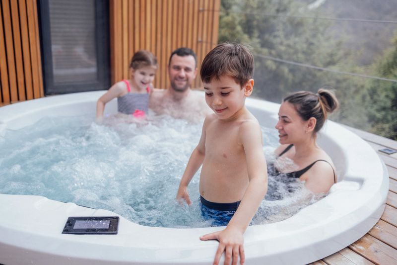 Happy family with two small children have fun in the hot tub. Cheerful young dad splashes water on his laughing cute son. Smiling man, woman, funny son and cute daughter relaxing in bubbling water