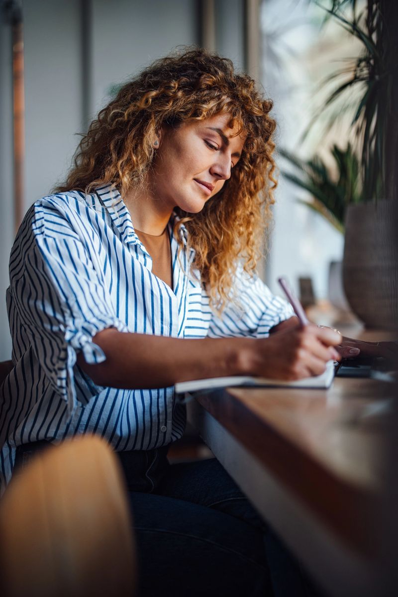 Young woman sitting in a cafe, writing in a notebook. She is focused on her work in a cozy, friendly environment.