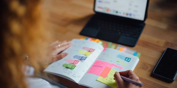 Person organizing tasks with colorful sticky notes in a planner beside a laptop and phone.