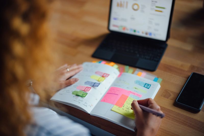 A person is planning and writing notes in a journal at a cozy cafe. A laptop and smartphone are on the desk.