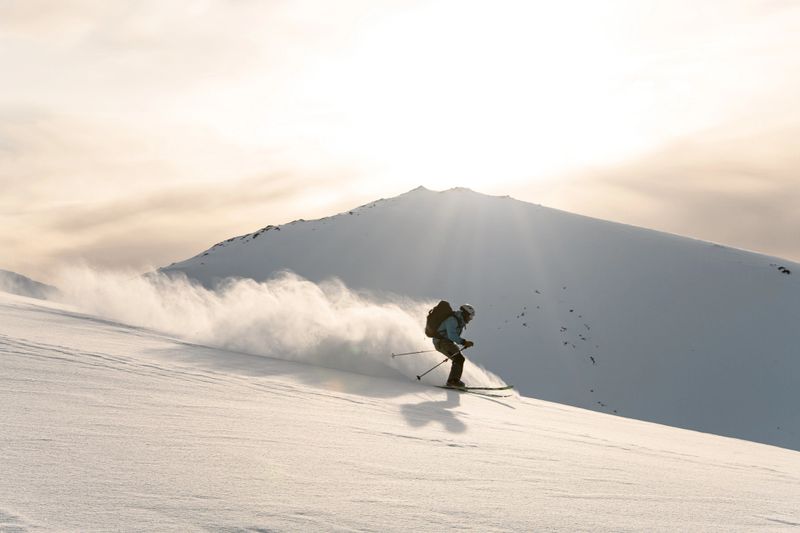 Extreme skier rushes down the mountain on skis, holding sticks for insurance in his hands, so that the powder of snow scatters behind him in different directions and behind him against the background of a bright sky on which the sun shines brightly
