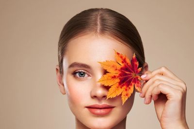 Young woman holding an autumn leaf over one eye, smiling gently.
