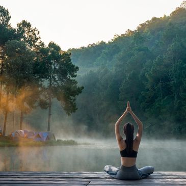 A woman practices yoga by a misty lake at sunrise in a forested area.