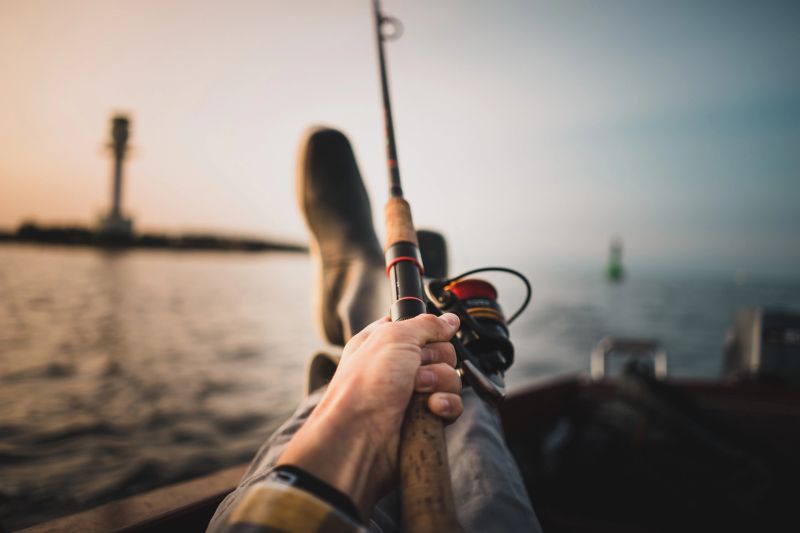 Low Section Of Man Fishing In Sea Against Sky During Sunset