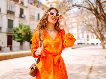 Smiling woman in orange dress and sunglasses walks outdoors on a sunny day.